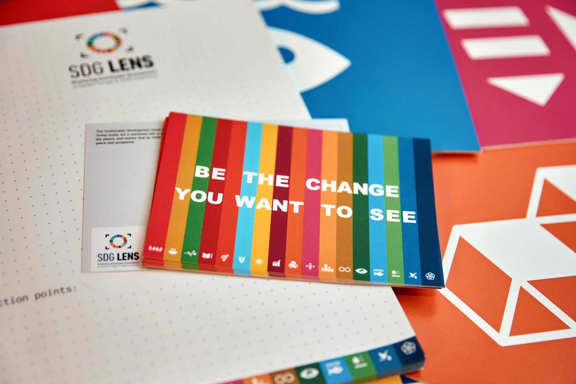 A booklet on the table, in SDG colors, the text on the cover: "Be the change you want to see". Everything is in SDG Lens brand colors.