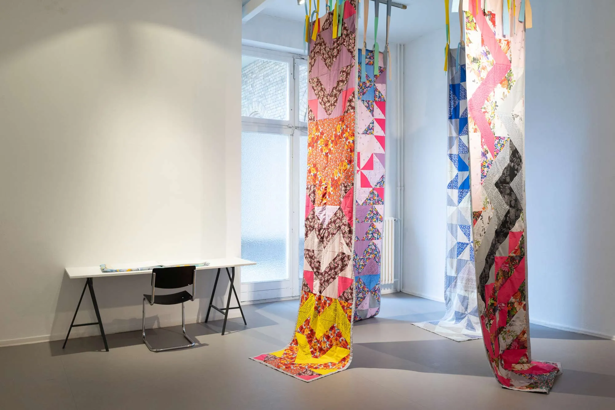 A photograph from the exhibition: a table and a chair with materials to read to the left, four patchwork sewn pieces of fabric hanging from the ceiling to the right