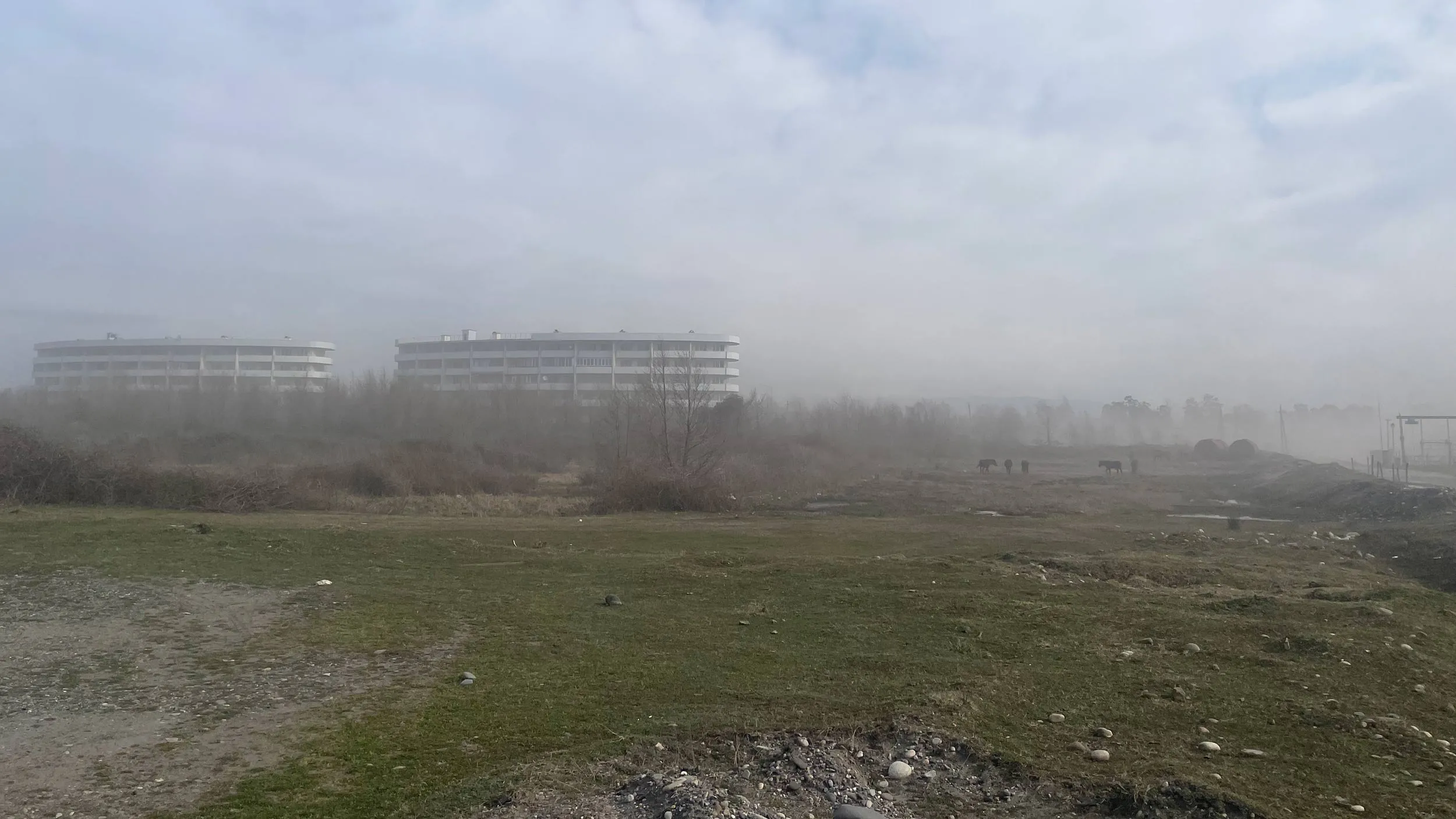 Two buildings standing in light fog, a wasteland in the foreground.