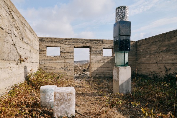 Artistic installation: a totem made of square- and tubular-shaped stone and minerals stacked on top of each other, located in an abandoned concrete building that has no roof.